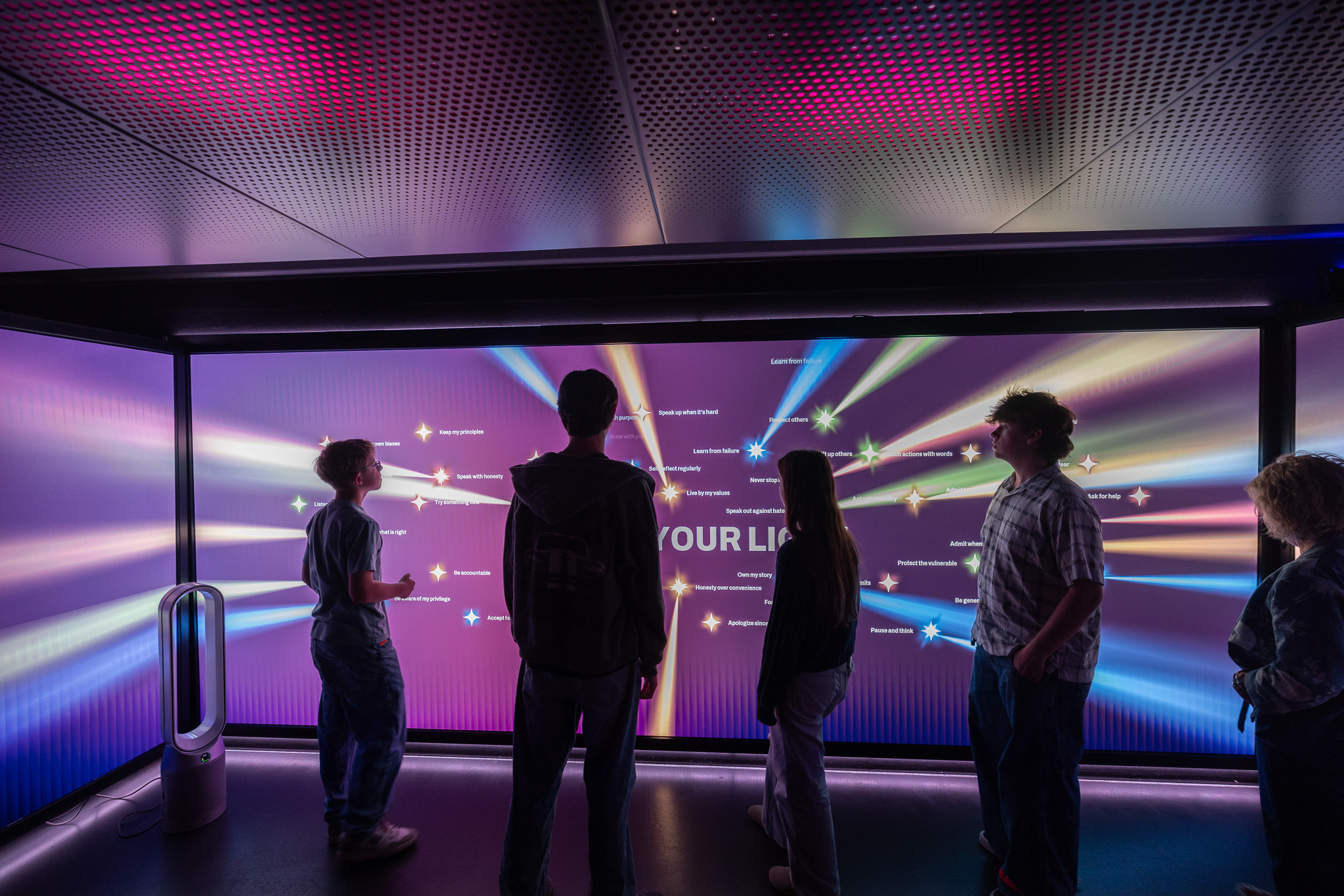 Students stand before the glowing Your Light wall of values inside Spark Interactive
