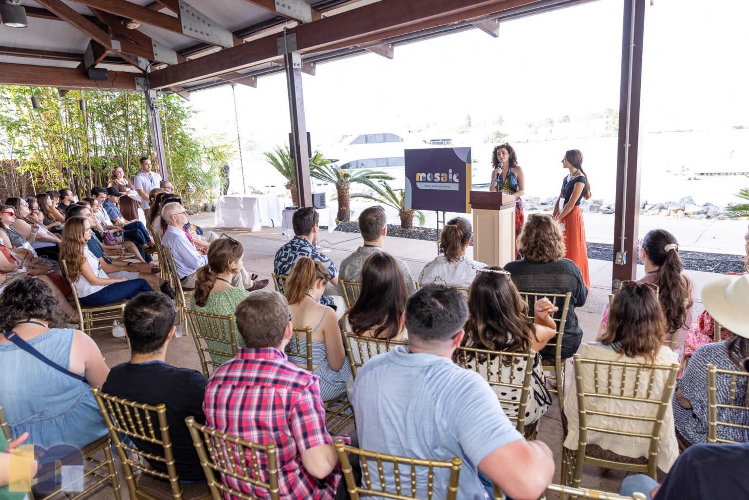 Members seated under a bayfront pavilion on Shelter Island, facing two speakers beside a Mosaic sign at the launch