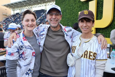 Three smiling people stand close together with arms around each other at a ballpark event. Two wear Padres caps and one wears a Padres pinstripe jersey, and a Southwest-sponsored Padres floral shirt is worn by one of them.