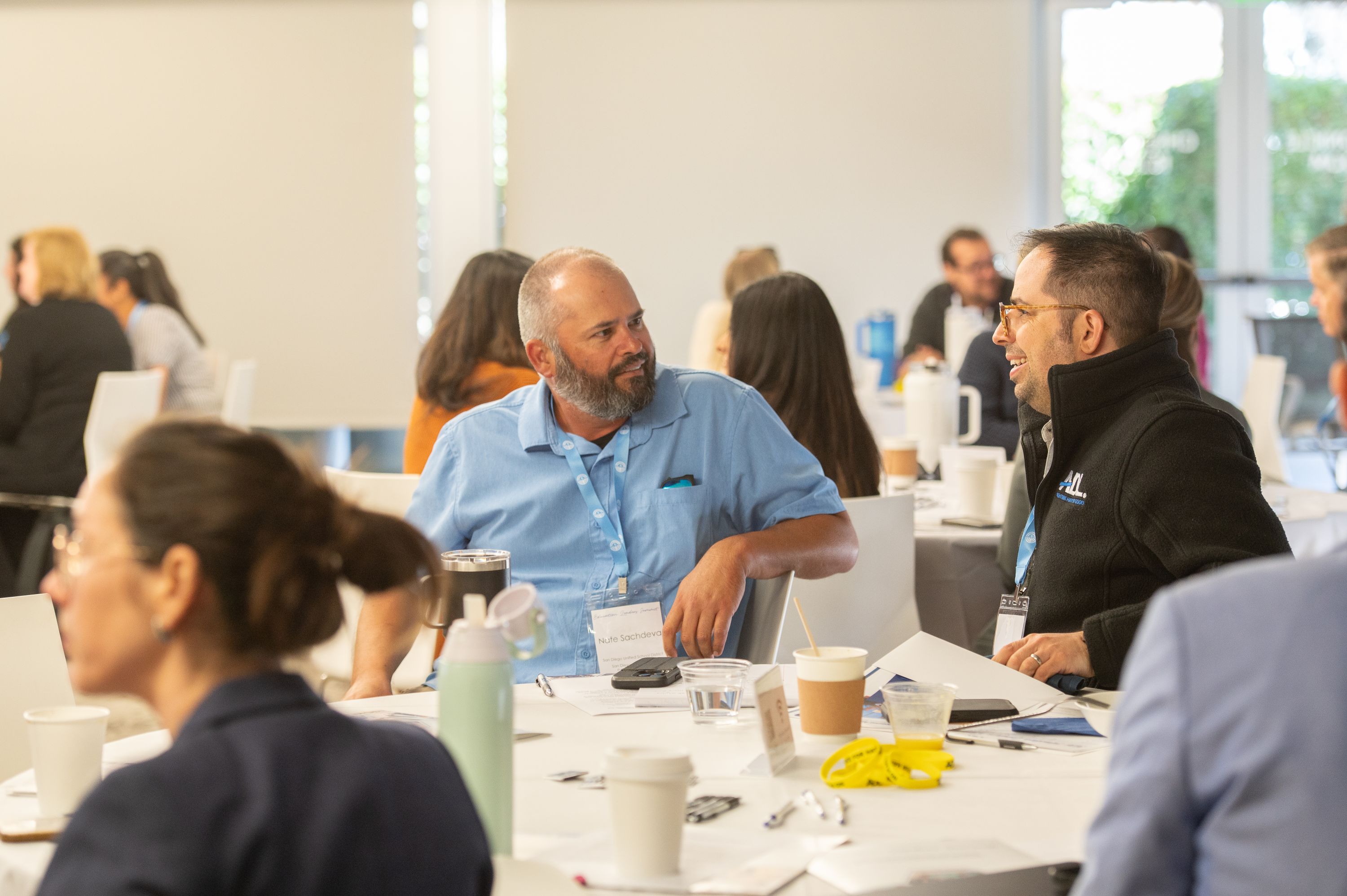 Several attendees sit around a white table in a bright meeting room, with coffee cups, water glasses and notebooks in front of them. Two men in the foreground are smiling and talking, while others gather and chat in the background.