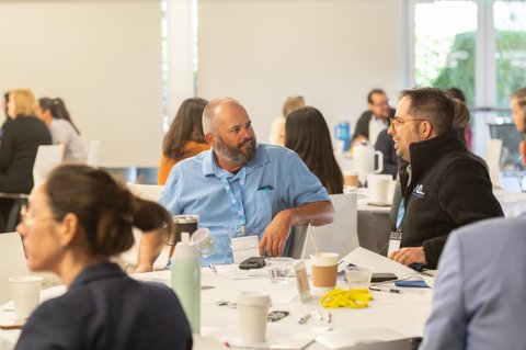 Several attendees sit around a white table in a bright meeting room, with coffee cups, water glasses and notebooks in front of them. Two men in the foreground are smiling and talking, while others gather and chat in the background.