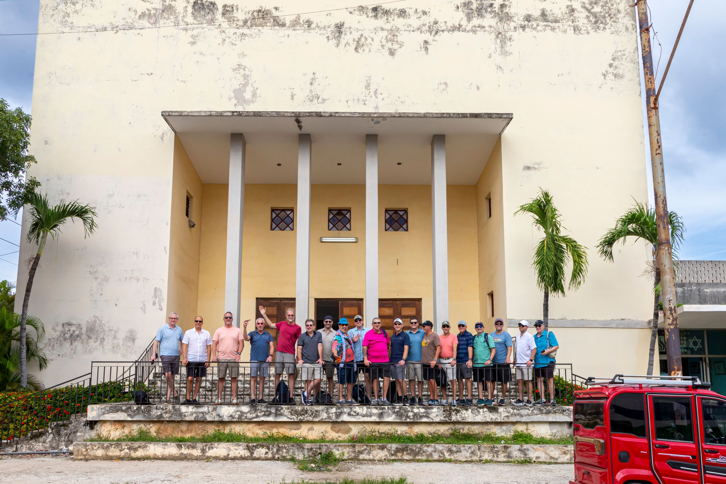 A group of about twenty men in casual summer clothes stands on the front steps of a pale yellow synagogue with white columns. A red van is parked at the right, and a Star of David is visible on a building at the far right.