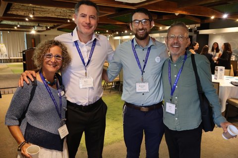 Four smiling adults pose arm in arm in a conference hall with wooden beamed ceiling and round tables behind them. All wear lanyards, and one carries a coffee cup.