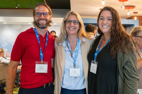 Three smiling people wearing blue lanyards stand close together and pose for the camera in a bright indoor venue. Two are wearing name badges, which are not transcribed here. Other attendees mill about in the background near tables and lounge seating.