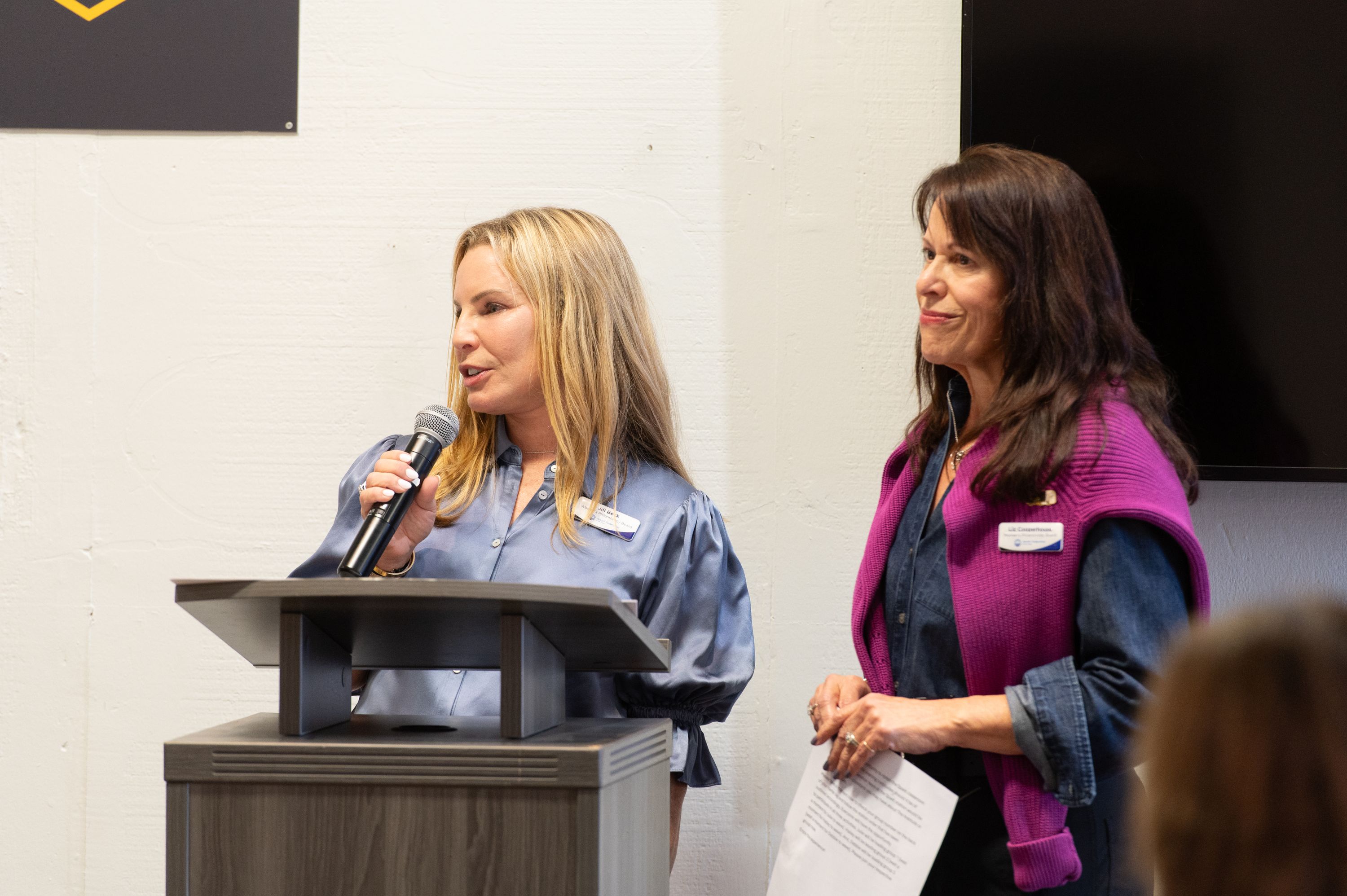 A woman in a blue satin blouse speaks into a microphone at a wooden podium while another woman in a magenta sweater and denim shirt stands beside her holding a printed page. A dark screen and a dark banner are visible on the wall behind them.