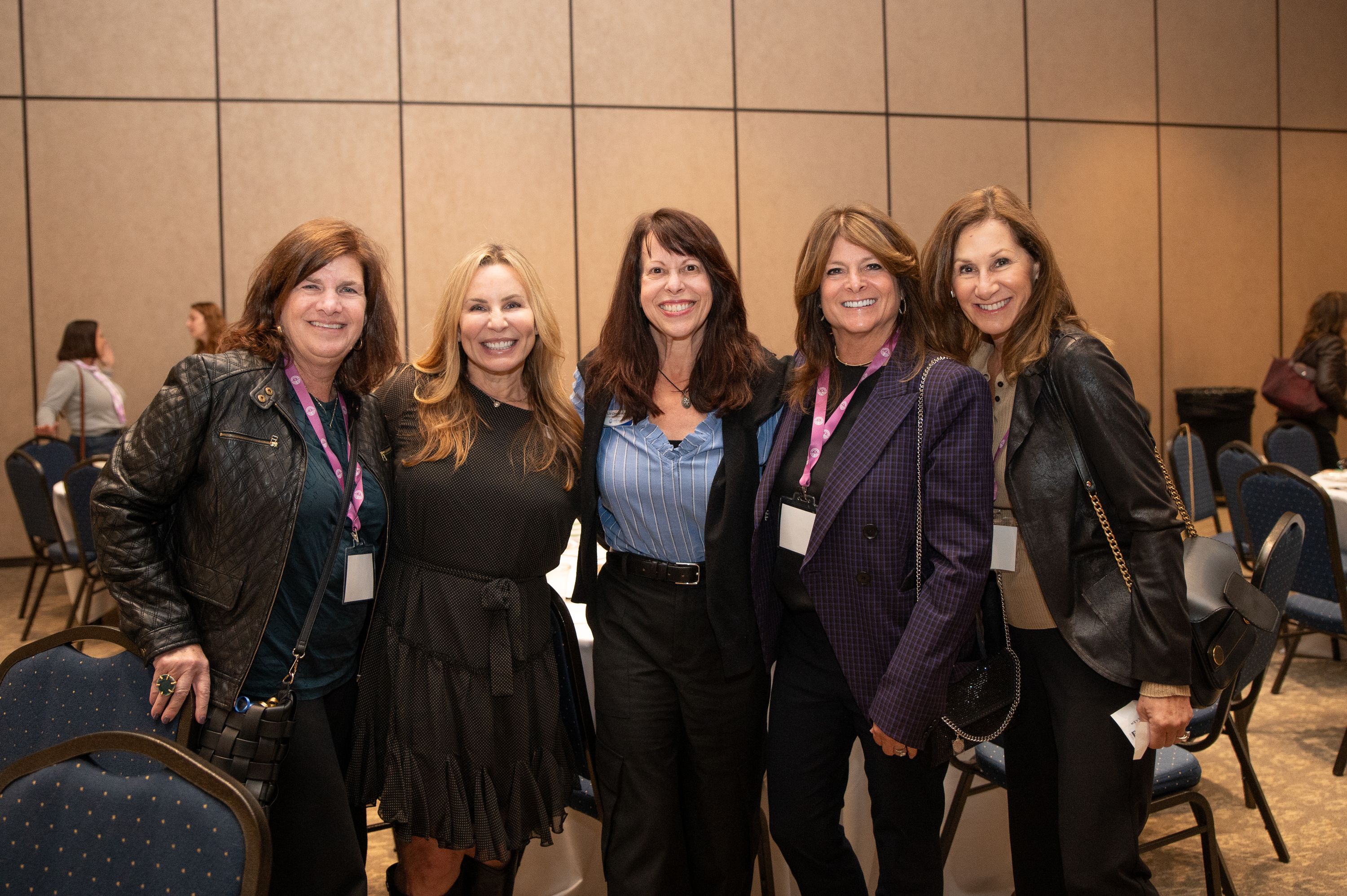 Five women pose arm in arm in a hotel ballroom with beige walls and blue banquet chairs. Several wear pink lanyards and name badges, which are not transcribed here.