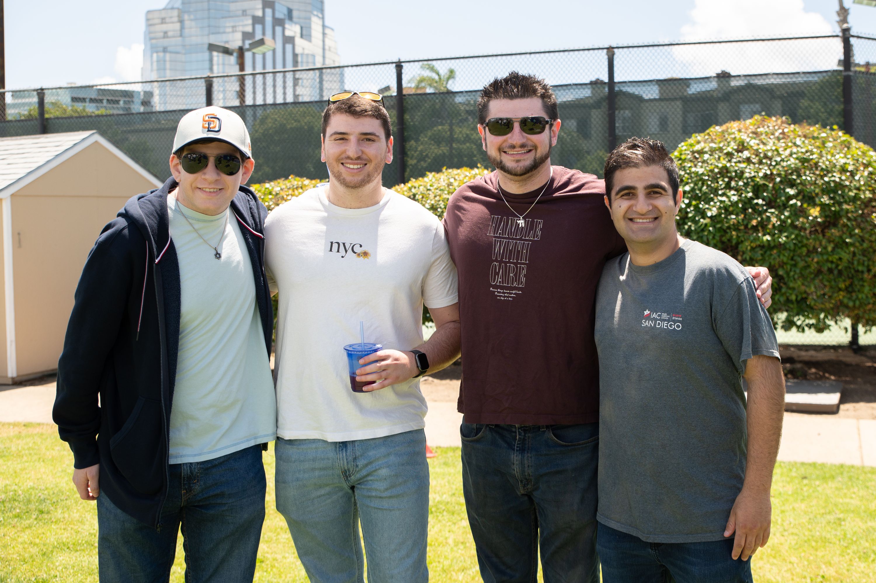 Four smiling men stand close together on a grassy lawn on a sunny day, with a tall hedge, a fence and city towers behind them. One wears a San Diego Padres cap and sunglasses, and another holds a blue drink cup.