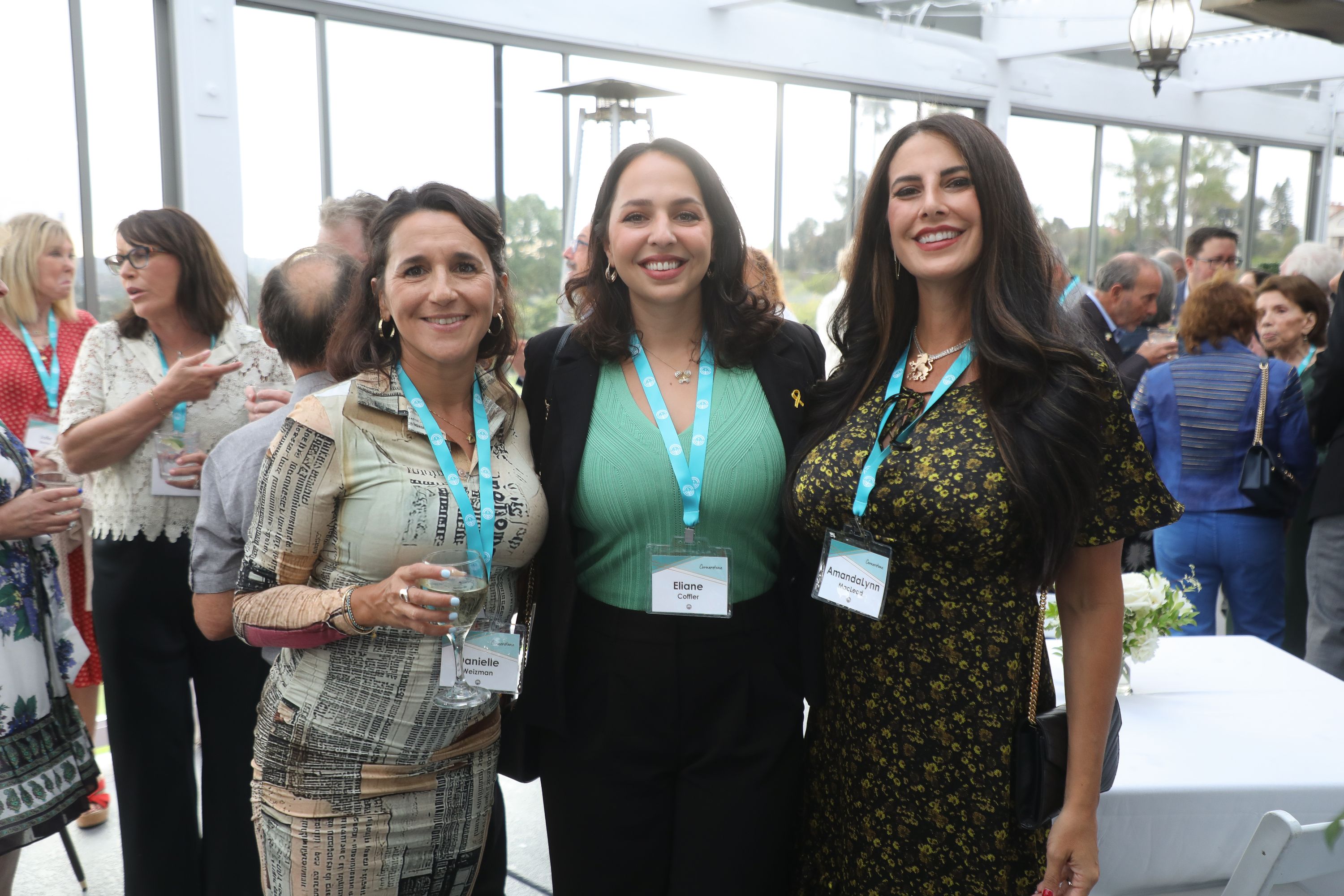 Three women pose together for a photo at an indoor reception with glass walls and greenery outside. Several of them wear teal lanyards, and two hold drinks.