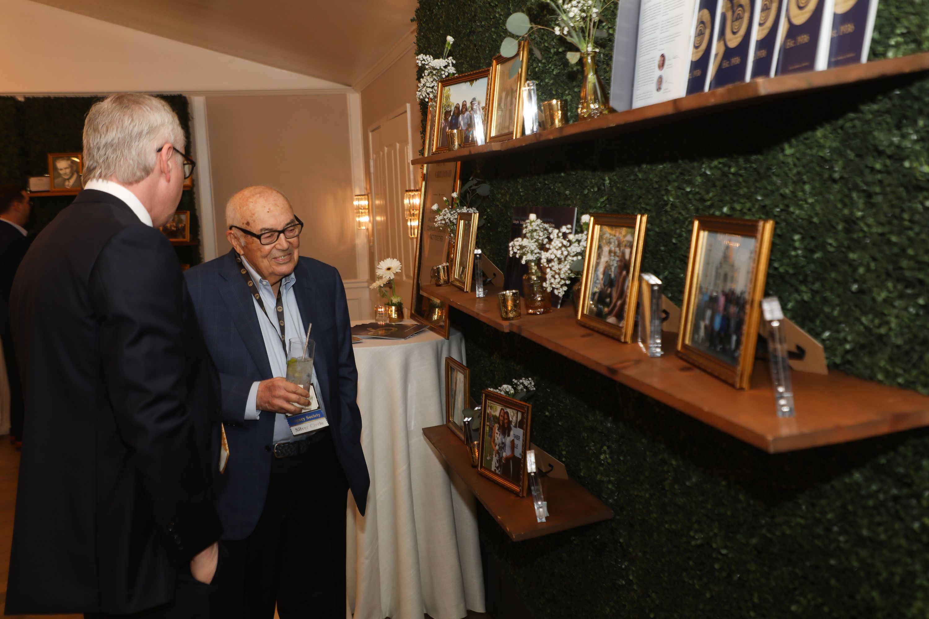 Two men stand in a room lined with green hedge panels and wooden shelves holding gold-framed photos, glass awards and small flower arrangements. An older man in a blue blazer and glasses holds a drink and looks at the displays while a man in a dark suit stands beside him.