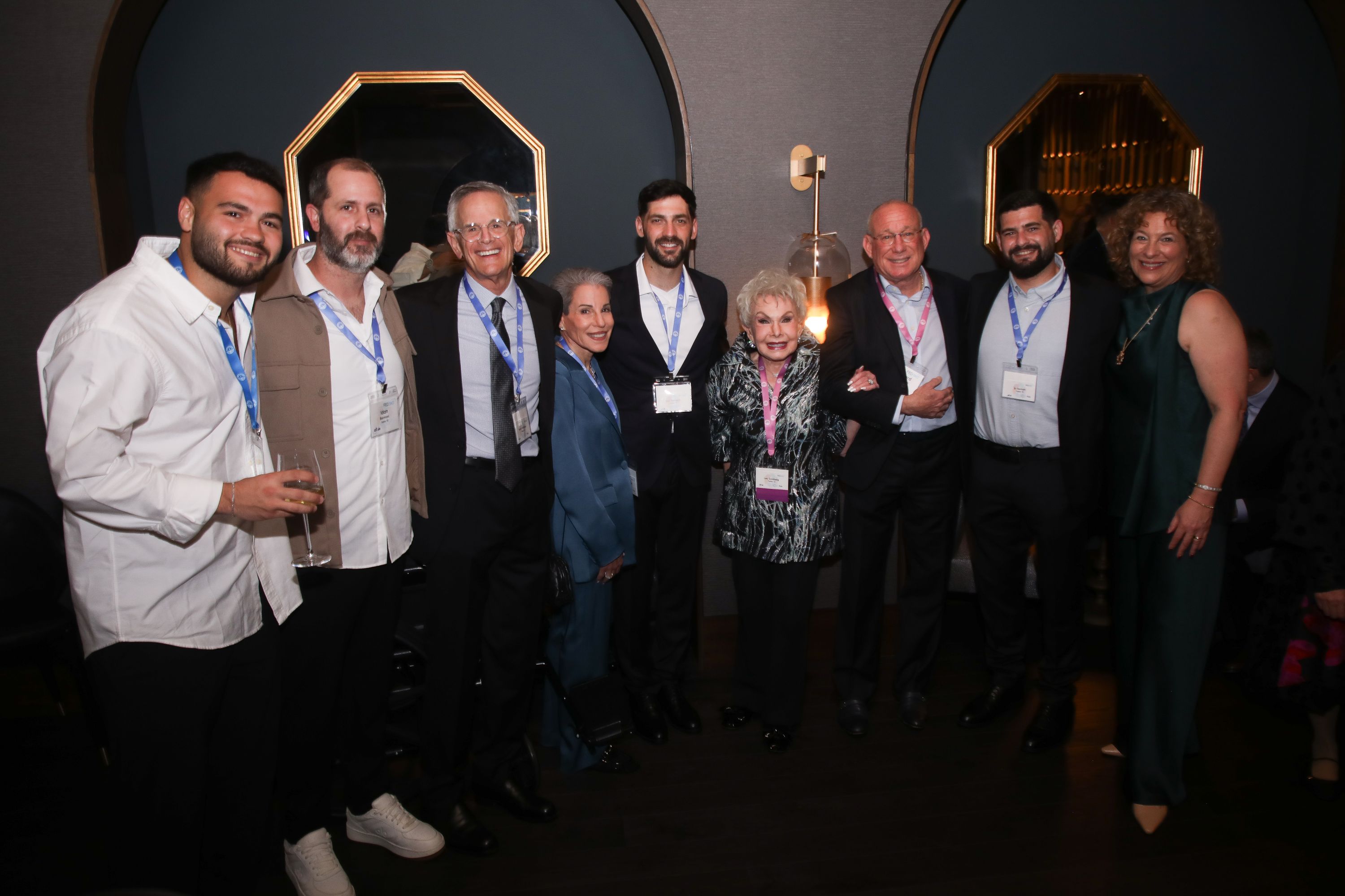 Nine guests pose together side by side in a dimly lit, elegant room with arched alcoves and gold-framed octagonal mirrors. Many wear conference lanyards and name tags, and one man holds a glass of wine.