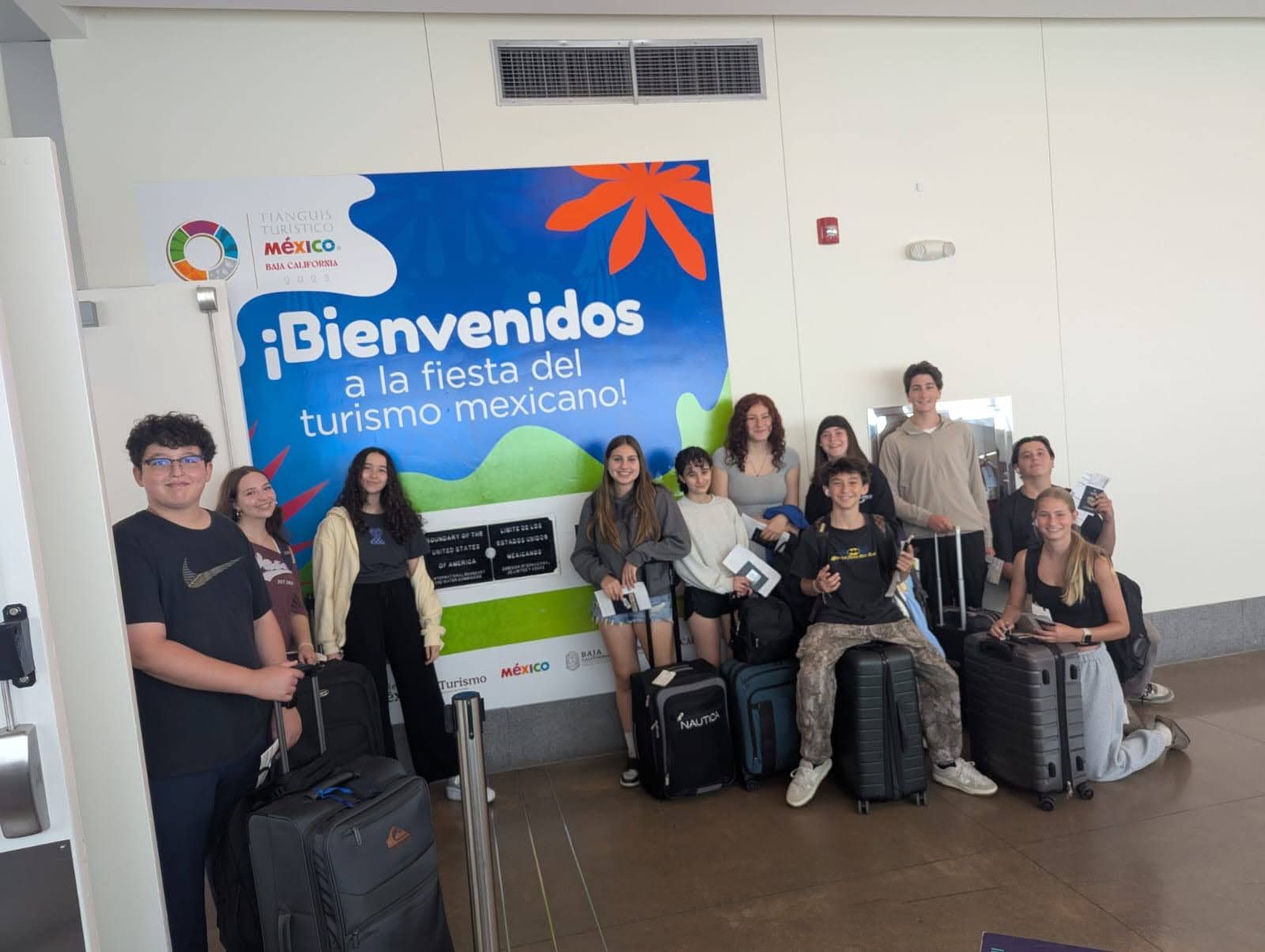 A group of teenagers stands and kneels together in front of a blue Mexican tourism welcome sign at an airport, with rolling suitcases around them. Some hold travel papers and phones, suggesting the start of a trip.