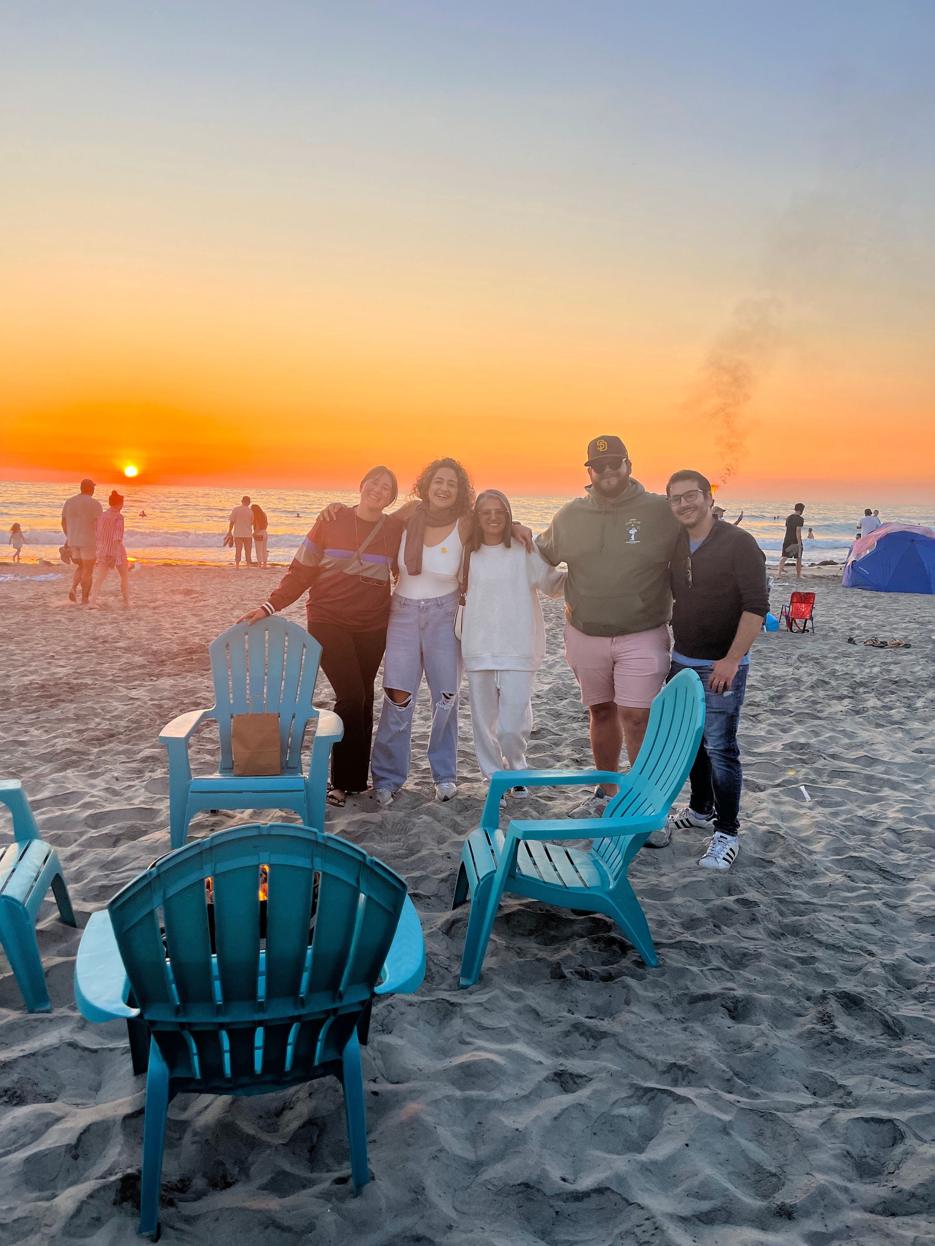 Five young adults stand arm in arm on a sandy beach in front of blue Adirondack chairs as the sun sets over the ocean. Other beachgoers and a blue tent are visible in the background.
