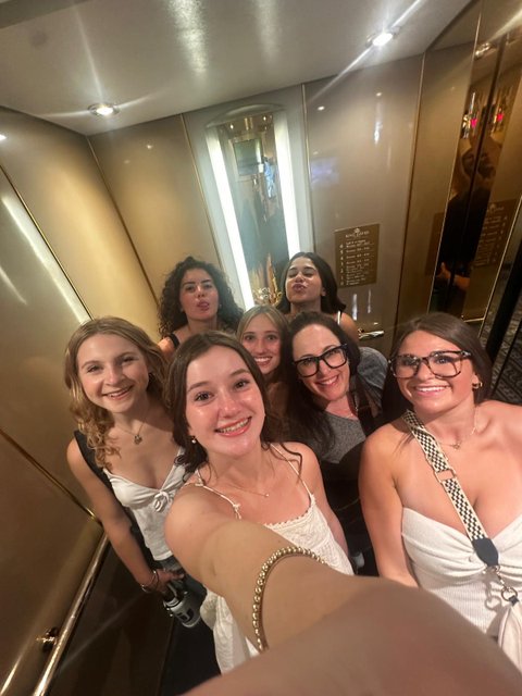 Seven women crowd into a gold-paneled elevator and pose for a selfie, several smiling and one pursing her lips. A floor directory reading King David is visible on the wall, suggesting a hotel stay.