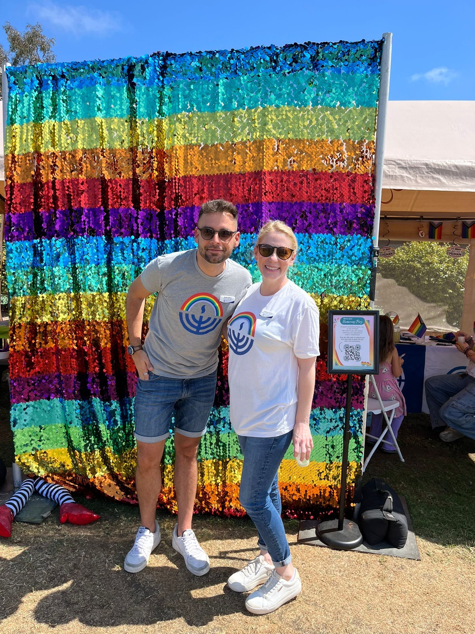 Two smiling adults in matching Jewish Federation-style shirts with a rainbow menorah logo pose in front of a shimmering rainbow sequin backdrop at an outdoor Pride event. A framed QR code sign stands beside them, and a tent and sunny lawn fill the background.