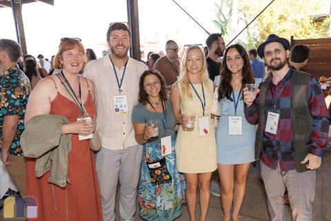 A group of six young adults poses together and smiles at an outdoor networking gathering, holding plastic cups and wearing conference lanyards. Some wear name badges, which are not transcribed.