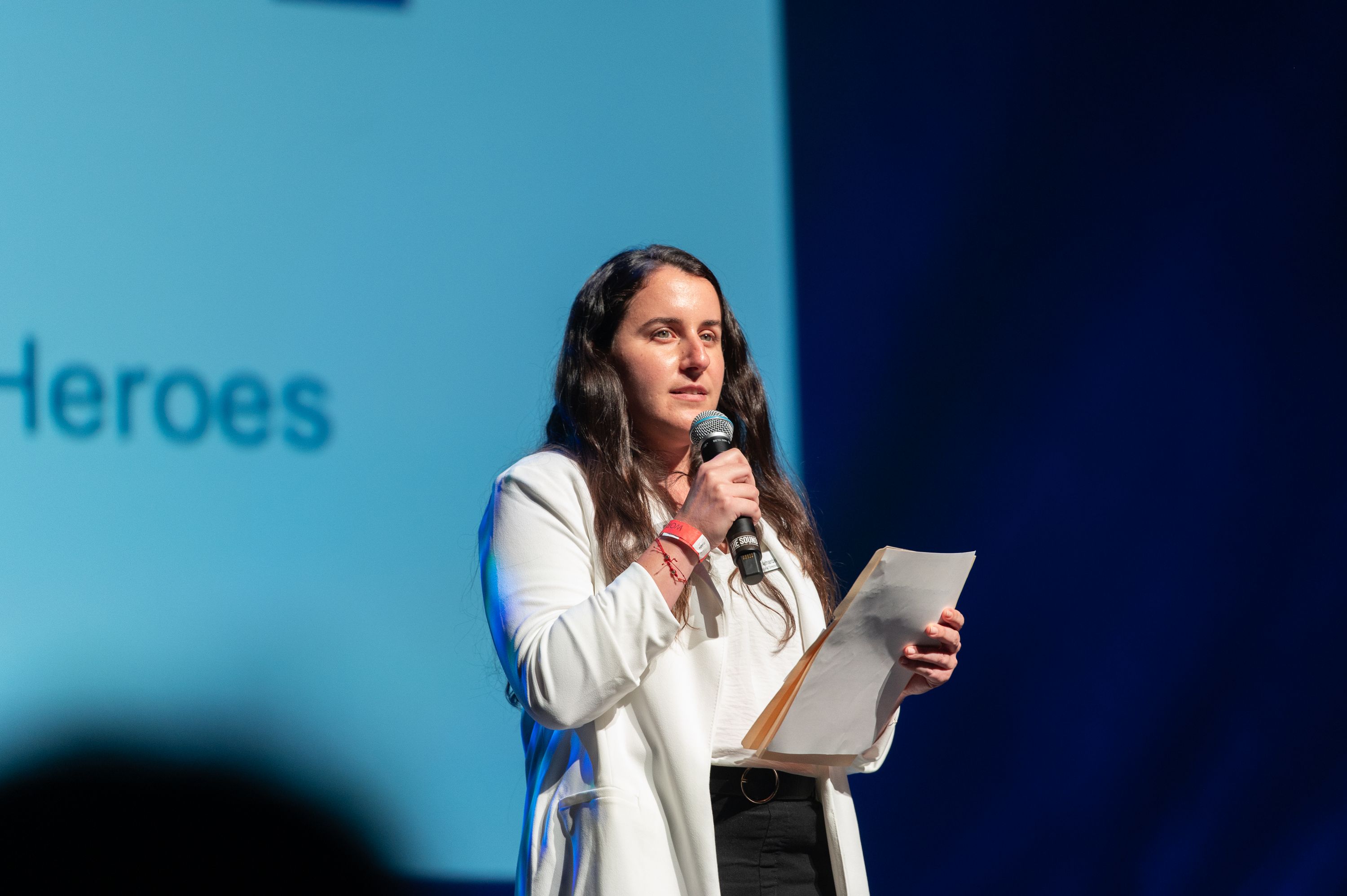 A woman in a white jacket stands on stage holding a microphone and a sheet of paper while speaking. Part of a blue screen behind her shows the word Heroes in large letters.