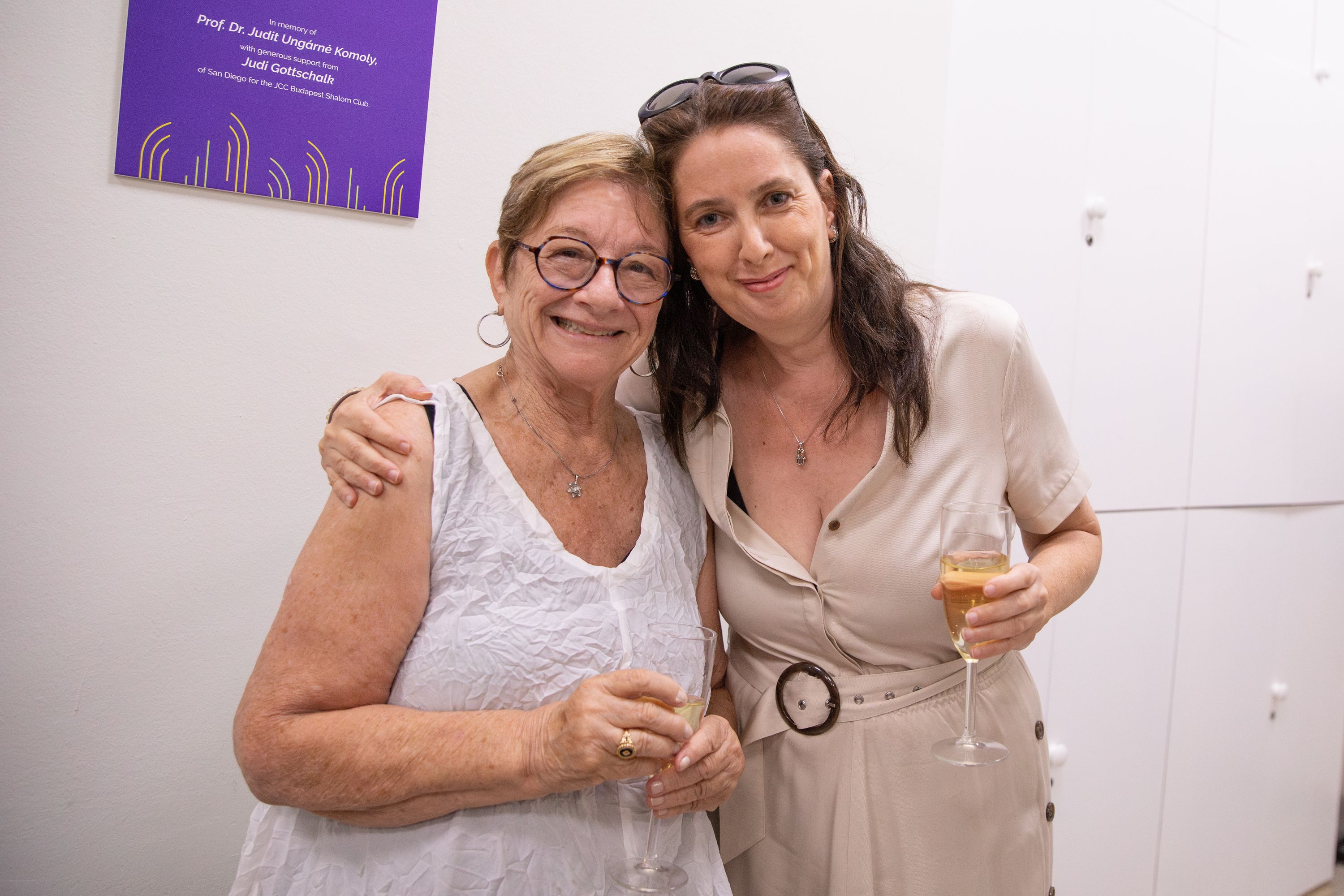 Two smiling women stand arm in arm, each holding a glass of sparkling wine, beside a purple plaque in memory of a professor. The plaque notes a gift for the JCC Budapest Shalom Club.