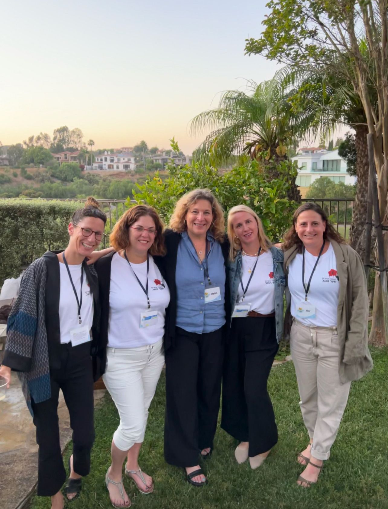 Five women in matching white t-shirts stand arm in arm on a grassy lawn at dusk, smiling for the camera. Palm trees and hillside homes are visible behind them, and each wears a name badge that is not transcribed.