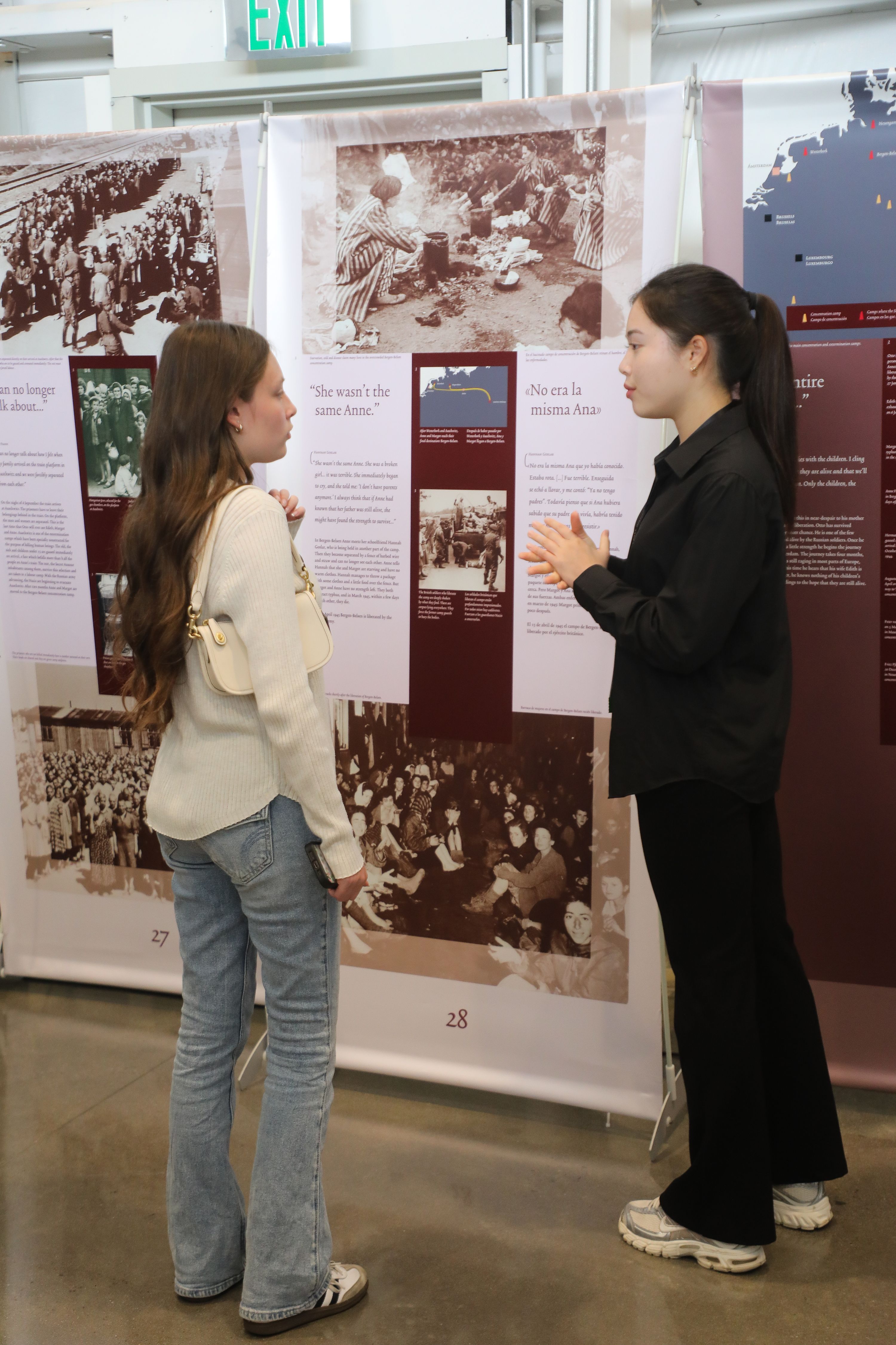 Two young women stand facing a multilingual Holocaust exhibition display with black-and-white historical photographs and quotations in English and Spanish. One gestures while speaking, and the other listens with her back partly to the camera.