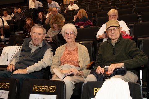 Three older adults smile for the camera from reserved seats in a large auditorium, with other attendees seated in rows behind them. Programs are visible in some hands, suggesting a community event such as a Yom HaShoah commemoration.
