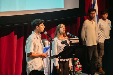 Two young people sing at microphones on a stage near a flag, with a candle and a floral arrangement at the podium. Two more young men stand behind them, one beside an Israeli flag, as a screen glows above the red curtains.